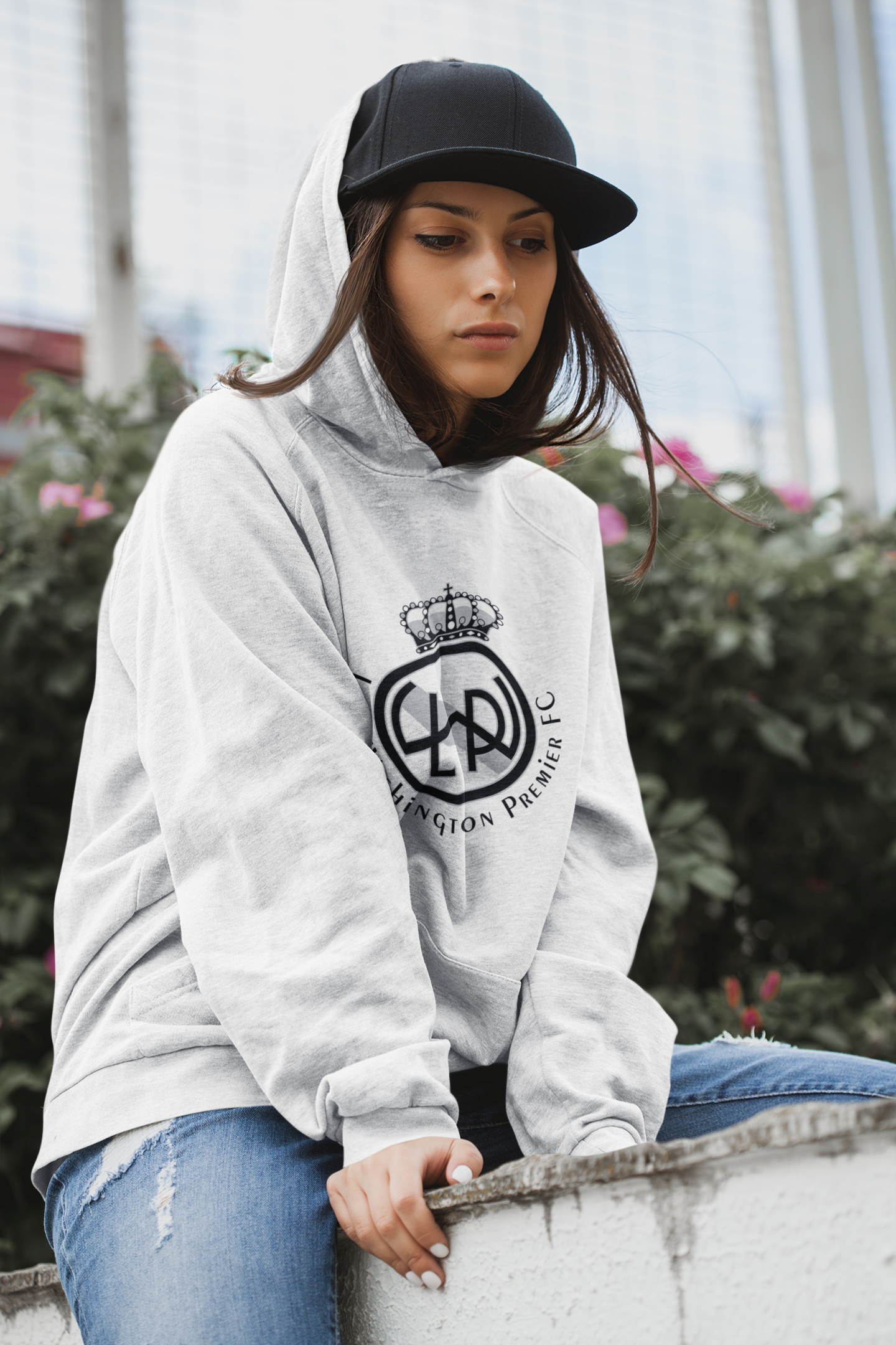 A young women sitting on a wall wearing a Lake Washington Premier FC light gray hoodie with the club crest in black and white on the chest. She is wearing a black cap with the hood up over her head and covering the cap