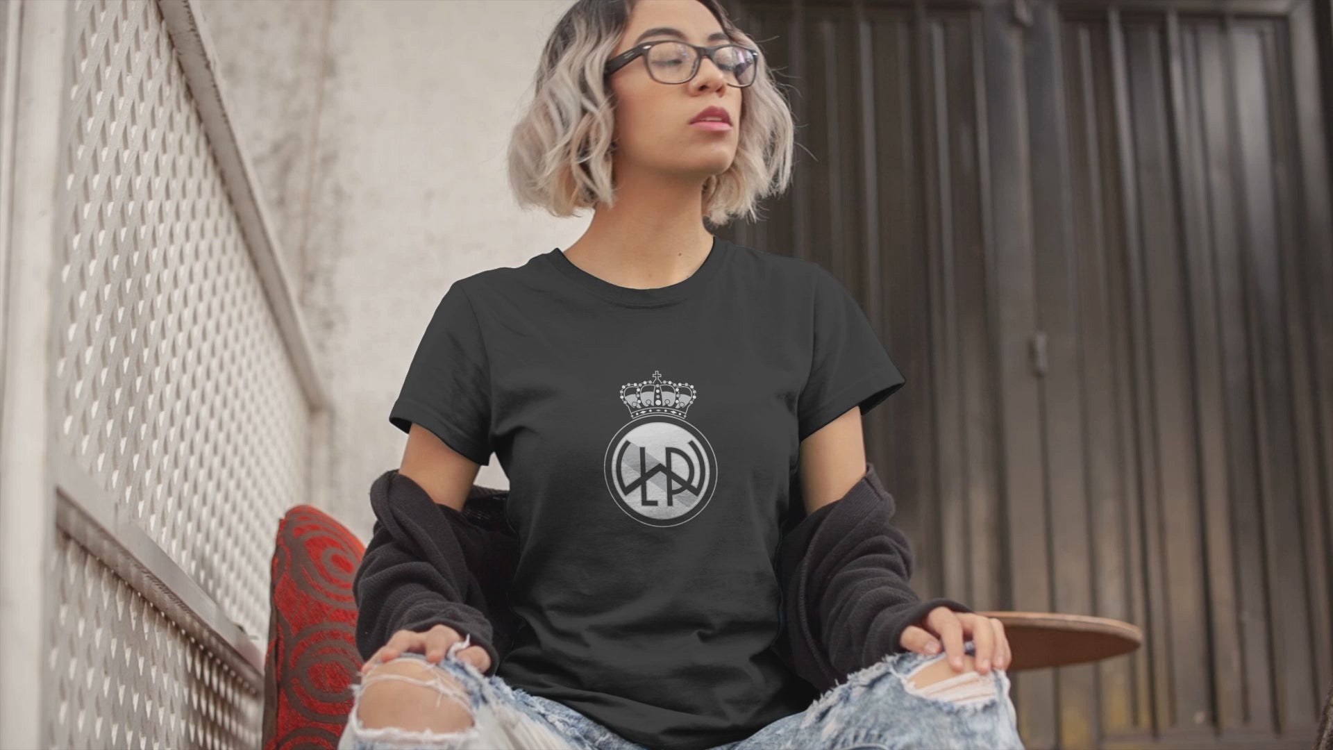 Load video: A t-shirt video featuring an edgy woman with blue framed glasses and short colored hair wearing an LWPFC dark gray short sleeved t-shirt with the club logo in black and white.